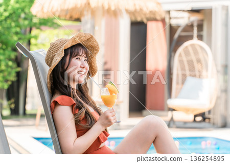 一位年輕的亞洲女子穿著泳裝，坐在泳池邊的躺椅上喝著熱帶果汁。 136254895