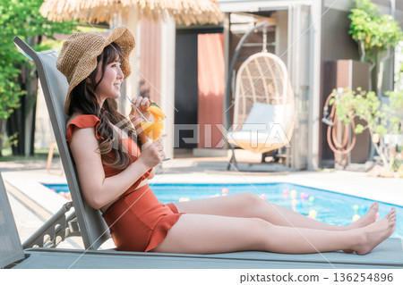 一位年輕的亞洲女子穿著泳裝，坐在泳池邊的躺椅上喝著熱帶果汁。 136254896