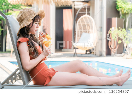 一位年輕的亞洲女子穿著泳裝,坐在泳池邊的躺椅上喝著熱帶果汁。 一位年輕的亞洲女子穿著泳裝,坐在泳池邊的躺椅上喝著熱帶果汁。 136254897