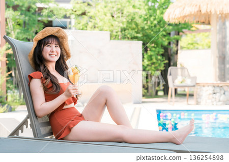 一位年輕的亞洲女子穿著泳裝,坐在泳池邊的躺椅上喝著熱帶果汁。 一位年輕的亞洲女子穿著泳裝,坐在泳池邊的躺椅上喝著熱帶果汁。 136254898