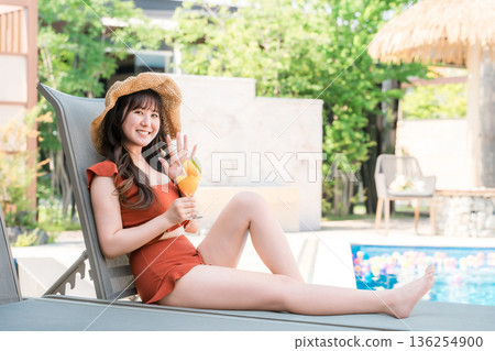 Young Asian woman in swimsuit sitting on recliner by pool and drinking tropical juice 136254900