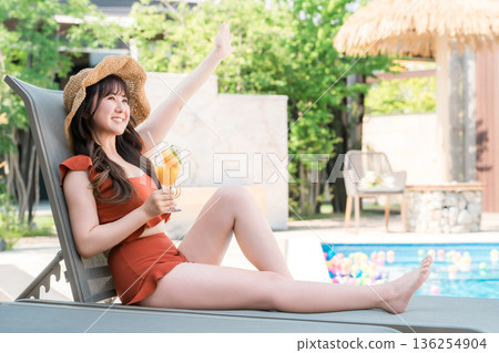 一位年輕的亞洲女子穿著泳裝,坐在泳池邊的躺椅上喝著熱帶果汁。 一位年輕的亞洲女子穿著泳裝,坐在泳池邊的躺椅上喝著熱帶果汁。 136254904