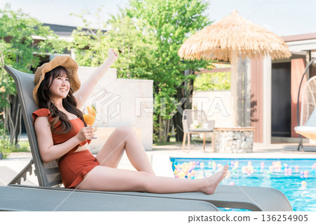 Young Asian woman in swimsuit sitting on recliner by pool and drinking tropical juice Young Asian woman in swimsuit sitting on recliner by pool and drinking tropical juice 136254905