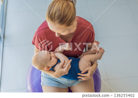 Young mother gently rocks her baby on a fitness ball at home, creating a calm loving atmosphere. Authentic family lifestyle, natural parenting and emotional bonding. Motherhood, wellbeing and baby Young mother gently rocks her baby on a fitness ball at home, creating a calm loving atmosphere. Authentic family lifestyle, natural parenting and emotional bonding. Motherhood, wellbeing and baby 136255402