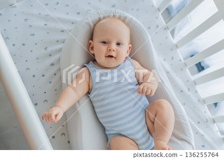 Baby lying peacefully in a cozy cocoon nest inside a crib, feeling safe and calm at home. Soft light and warm atmosphere highlight comfort and care. Infant sleep, safety and gentle parenting concept 136255764