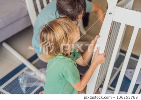 Father and older brother assembling a baby crib at home, preparing for a newborn arrival. Warm family moment full of care and anticipation. Parenthood, sibling bond and home preparation concept 136255769