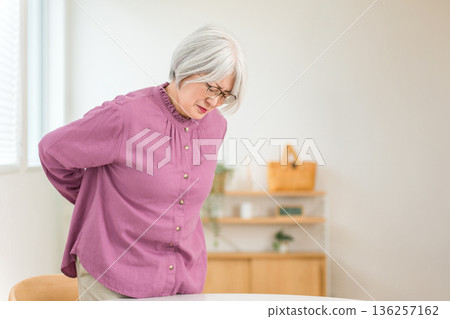 A gray-haired senior woman who seems to be suffering from lower back pain and holding her lower back A gray-haired senior woman who seems to be suffering from lower back pain and holding her lower back 136257162