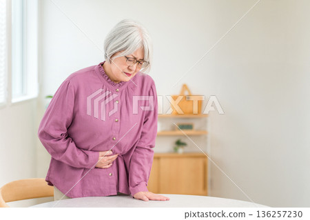 A gray-haired senior woman with a painful expression holding her stomach due to stomach pain 136257230