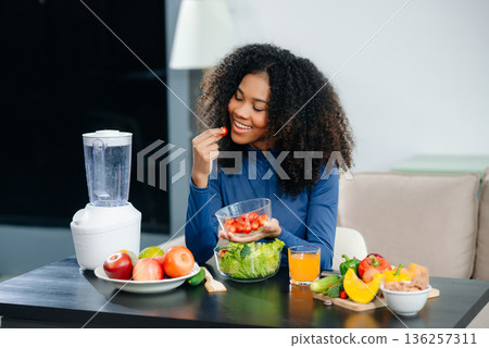 woman enjoys cherry tomatoes and salad in a cozy home kitchen surrounded by fruits, juice, and a blender. Perfect for health, diet, and clean eating concepts. 136257311