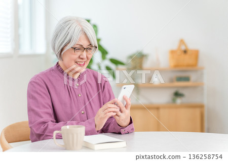一位老婦人坐在餐桌旁使用智慧型手機(面帶微笑,看起來很開心) 一位老婦人坐在餐桌旁使用智慧型手機(面帶微笑,看起來很開心) 136258754