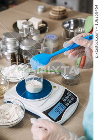 Person wearing gloves uses blue spatula to add white powder into glass beaker on digital scale. micro-batch skincare, handcrafted beauty ritual, botanical alchemy, small business aesthetic. Person wearing gloves uses blue spatula to add white powder into glass beaker on digital scale. micro-batch skincare, handcrafted beauty ritual, botanical alchemy, small business aesthetic. 136259454