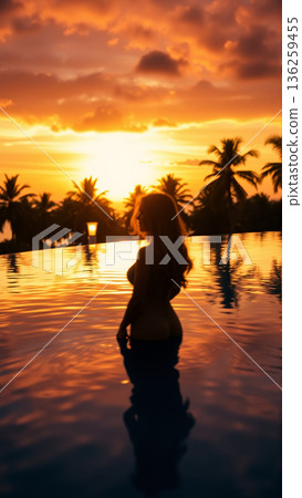 Silhouette of a beautiful woman relaxing in the pool in the setting light of the sun. The pool is surrounded by tropical trees. 136259455