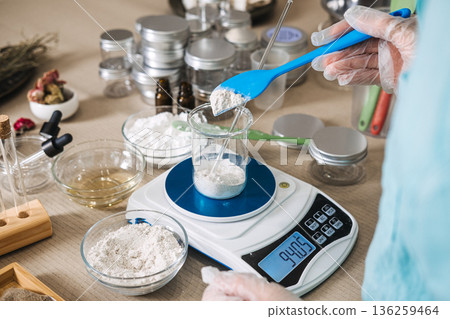 Person wearing gloves uses blue spatula to add white powder into glass beaker on digital scale. micro-batch skincare, handcrafted beauty ritual, botanical alchemy, small business aesthetic. Person wearing gloves uses blue spatula to add white powder into glass beaker on digital scale. micro-batch skincare, handcrafted beauty ritual, botanical alchemy, small business aesthetic. 136259464