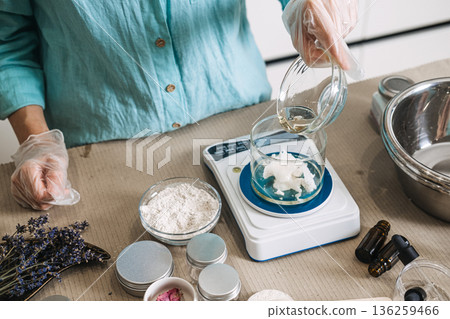 Hand in glove pours liquid oil from small bowl into jar containing white solid butter on digital scale. raw botanical oils, cold pressed skincare, nutrient dense beauty, unrefined ingredients. 136259466