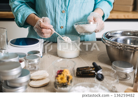 Person wearing gloves adds powder to liquid mixture during artisanal skincare formulation. plastic-free beauty making, sustainable cosmetic lab, eco-conscious diy, bulk ingredient aesthetic. 136259482
