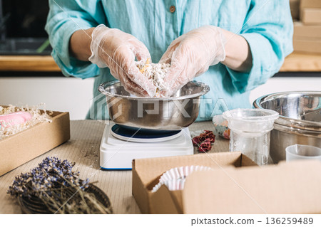 Person mixes botanical ingredients with hands in metal bowl on digital scale surrounded by packaging materials. Clean beauty process, transparent skincare brand, handmade cosmetic workshop 136259489