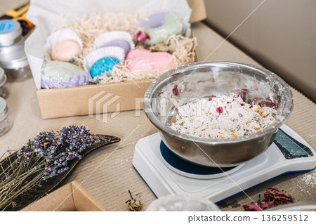 Metal bowl on digital scale contains white powder mixture with colorful flower petals next to gift box of bath bombs. Botanical bath products, dried flowers, natural wellness, flower petal bath. 136259512