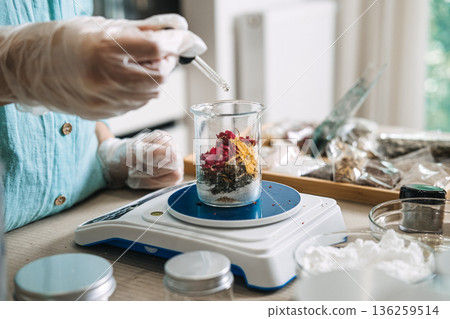 Person uses pipette to add liquid into glass beaker containing dried flowers on digital scale. home lab, precision DIY beauty, aesthetic apothecary equipment, scientific handmade skincare. 136259514