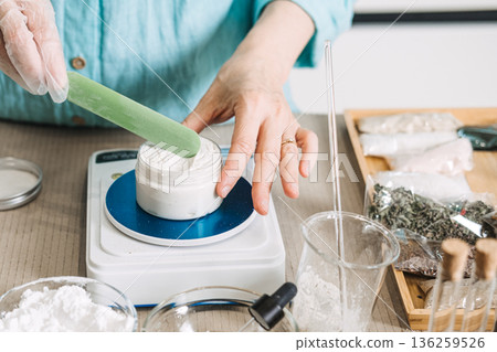 Hands use green spatula to mix white cream in jar on digital scale. Natural skincare formulation, chemical-free cosmetics, botanical ingredients, non-toxic beauty. Hands use green spatula to mix white cream in jar on digital scale. Natural skincare formulation, chemical-free cosmetics, botanical ingredients, non-toxic beauty. 136259526