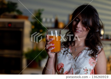 Woman holds glowing beverage joyfully, Caucasian female enjoying bright juice in kitchen, Amber liquid radiates warmth while woman gazes with subtle happiness in background 136260919