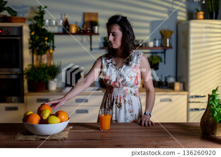 Female picks fruit from counter, Woman in kitchen selects fresh oranges, Female in comfortable kitchen environment extends hand to pick ripe oranges from wooden tabletop Female picks fruit from counter, Woman in kitchen selects fresh oranges, Female in comfortable kitchen environment extends hand to pick ripe oranges from wooden tabletop 136260920
