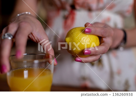 Hand holding lemon above drink, Closeup of lemon and hand preparing juice, Natural light illuminates hand and lemon during everyday juice preparation in cozy kitchen Hand holding lemon above drink, Closeup of lemon and hand preparing juice, Natural light illuminates hand and lemon during everyday juice preparation in cozy kitchen 136260938