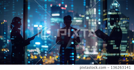Three business people stand together holding documents in a modern office. They are surrounded by a vibrant city skyline at night with many lights 136263141