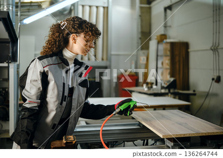 Carpenter works on wooden plank in workshop using spray gun to apply finish or sealant 136264744