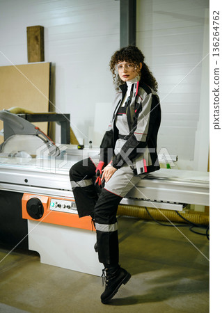 Woman in work clothes sits on a machine in a workshop during daytime while looking at the camera Woman in work clothes sits on a machine in a workshop during daytime while looking at the camera 136264762