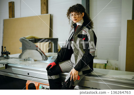 Woman in work outfit sits on a machine in a workshop during the day while wearing gloves Woman in work outfit sits on a machine in a workshop during the day while wearing gloves 136264763