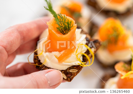 A gourmet smoked salmon canape elegantly arranged on a crispy cracker, topped with a swirl of smooth cream cheese, fresh thyme, and a drizzle of olive oil. The vibrant orange salmon contrasts A gourmet smoked salmon canape elegantly arranged on a crispy cracker, topped with a swirl of smooth cream cheese, fresh thyme, and a drizzle of olive oil. The vibrant orange salmon contrasts 136265460