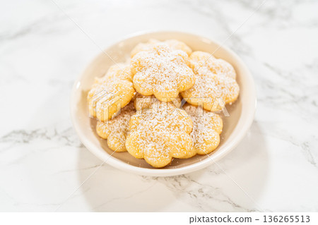 A small white bowl filled with freshly baked lemon spritz cookies, dusted with powdered sugar, sits on a marble countertop. These buttery, melt-in-your-mouth cookies have a delicate texture and a A small white bowl filled with freshly baked lemon spritz cookies, dusted with powdered sugar, sits on a marble countertop. These buttery, melt-in-your-mouth cookies have a delicate texture and a 136265513