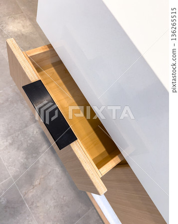 A close up of an open bathroom drawer shows light wood construction and a modern black handle. The drawer interior contrasts with the white countertop above for a sleek, minimal style. 136265515