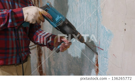 Chipping a wall with a hammer drill in a room undergoing renovation, a worker in brown pants and a checkered shirt holds a jackhammer and chisels a vertical hole in a shabby wall, chiseling a channel Chipping a wall with a hammer drill in a room undergoing renovation, a worker in brown pants and a checkered shirt holds a jackhammer and chisels a vertical hole in a shabby wall, chiseling a channel 136265772