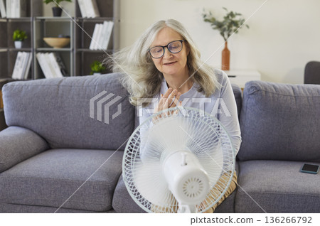 Senior woman feeling warm cooling herself with fan, room climate problem, uncomfortable to relief Senior woman feeling warm cooling herself with fan, room climate problem, uncomfortable to relief 136266792