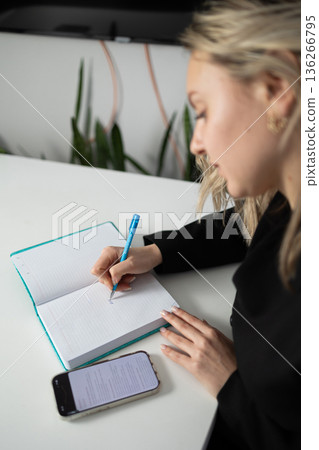 Business Woman Writes in Diary While Sitting at a Desk in an Office Setting Near Plants Business Woman Writes in Diary While Sitting at a Desk in an Office Setting Near Plants 136266795