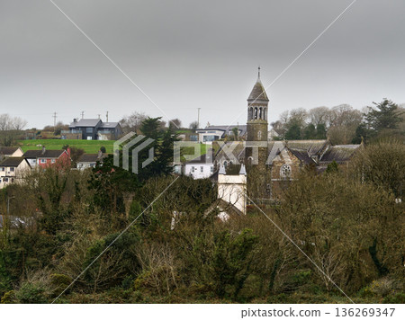 Historic stone church with bell tower stands among village houses, partially hidden by bare deciduous trees. A modern home sits on a green hill under a grey winter sky in rural Ireland. Historic stone church with bell tower stands among village houses, partially hidden by bare deciduous trees. A modern home sits on a green hill under a grey winter sky in rural Ireland. 136269347