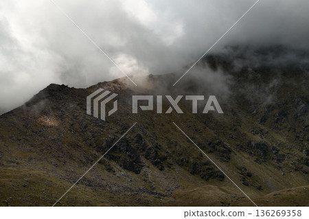 Steep, rugged mountain slopes ascend towards obscured jagged peaks. Heavy, grey clouds roll across the summit, creating a dramatic and misty atmosphere over the sparse, rocky terrain. Steep, rugged mountain slopes ascend towards obscured jagged peaks. Heavy, grey clouds roll across the summit, creating a dramatic and misty atmosphere over the sparse, rocky terrain. 136269358