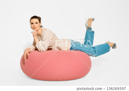 Model poses on pink bean bag while wearing casual clothes and high heels in studio setting Model poses on pink bean bag while wearing casual clothes and high heels in studio setting 136269397