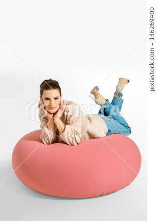 Woman poses on a large pink cushion while wearing jeans and a light sweater in a studio setting Woman poses on a large pink cushion while wearing jeans and a light sweater in a studio setting 136269400