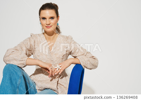 Woman sitting in a chair wearing a light top and jeans with a floral accessory on her hand Woman sitting in a chair wearing a light top and jeans with a floral accessory on her hand 136269408