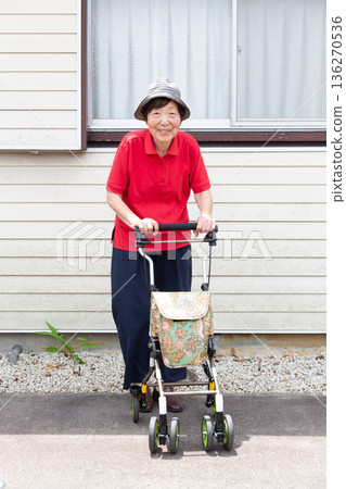 一位老年婦女站在戶外，雙手扶著一輛銀色推車，正面照，全身照。 136270536