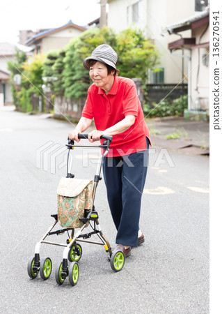 Senior woman walking across the road using a silver cart outdoors. Wheelchair. Whole body. Senior woman walking across the road using a silver cart outdoors. Wheelchair. Whole body. 136270541