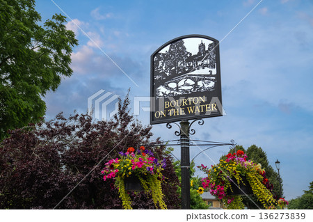 A tranquil roadside scene where an ornate village sign Bourton-on-the-Water, stands among blooming baskets and evening skies, looping softly through calm air. 136273839
