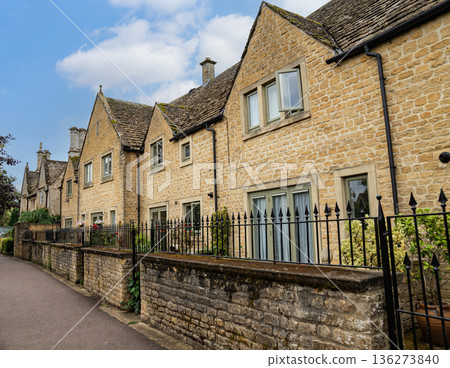 Stone houses with pitched roofs line a quiet path under a clear blue sky showing warm tones and timeless village charm in soft daylight. 136273840