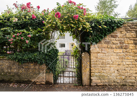 A floral archway of climbing roses and greenery frames an ornate iron garden gate,a bright country house beyond the stone wall. 136273845