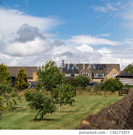 Houses with rooftop solar panels sit in a quiet countryside landscape beside an open orchard field under bright daylight. 136273855