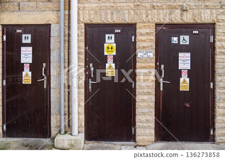 Outdoor public restroom doors on a stone building wall feature dark wooden panels with safety signs restroom icons and caution labels in bright daylight. 136273858