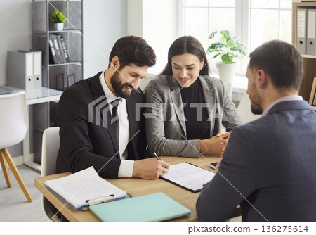 Happy young couple signing contract with male manager while reaching agreement in modern office. Happy young couple signing contract with male manager while reaching agreement in modern office. 136275614