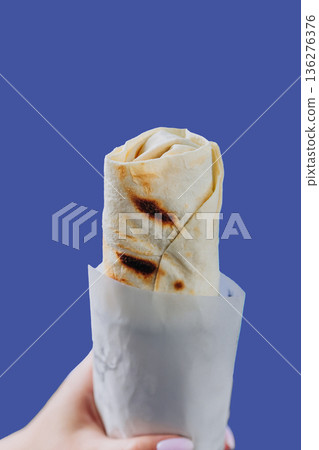 Hand holding wrapped shawarma in paper sleeve isolated on blue background Hand holding wrapped shawarma in paper sleeve isolated on blue background 136276376